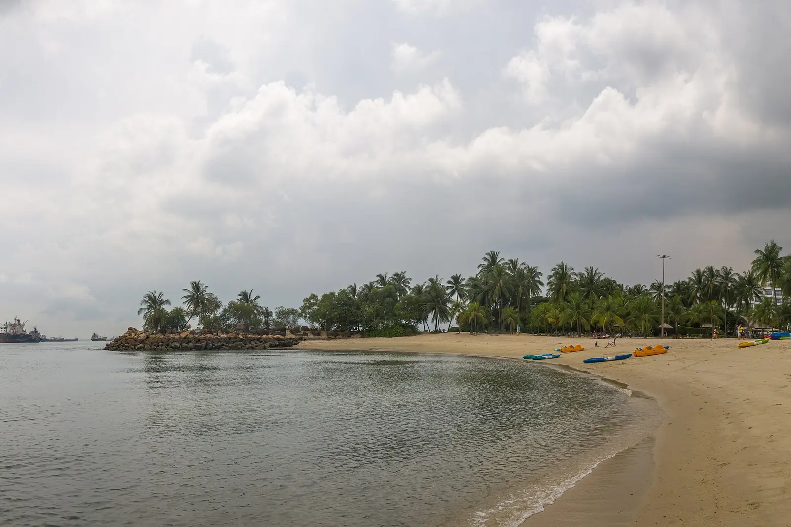 Un'ampia vista della spiaggia di Siloso sotto un cielo grigio, con la sabbia che si incurva lungo acqua calma e piccoli isolotti coperti di alberi