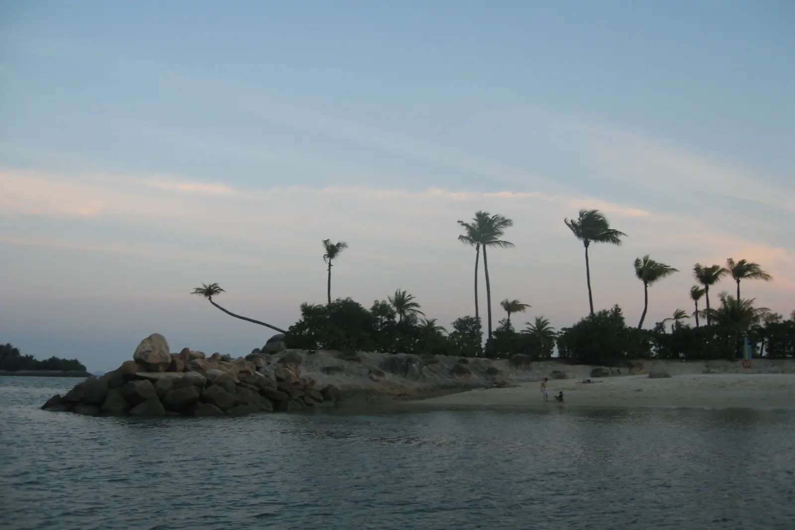 La spiaggia di Siloso al crepuscolo, con palme lungo un promontorio roccioso e un cielo rosa pallido e azzurro sull'acqua