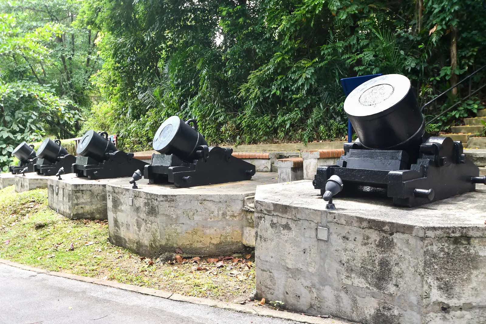 Una fila di mortai da trincea neri da 13 pollici conservati su basamenti di cemento tra gli alberi a Fort Siloso, a Sentosa