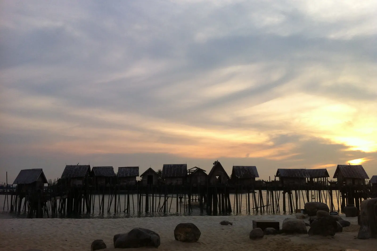 Il palco di Songs of the Sea, lo spettacolo sostituito da Wings of Time, con una fila di capanne su palafitte in silhouette contro il cielo al tramonto sulla spiaggia di Siloso